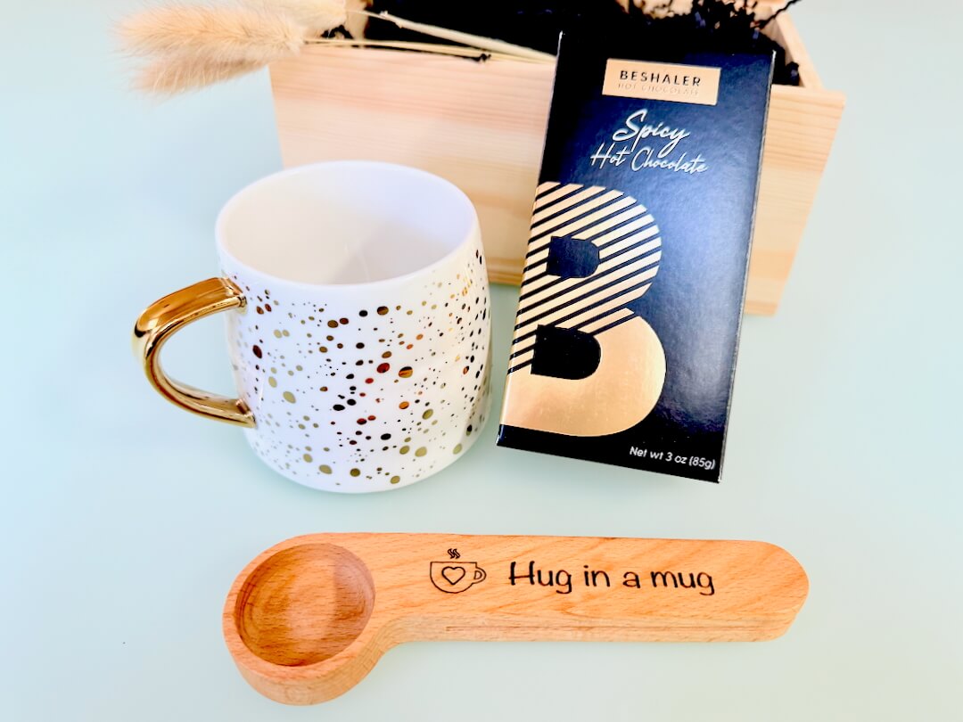 Golden dots mug, box of spicy hot chocolate, and wooden spoon with 'Hug in a mug' text on a light blue background